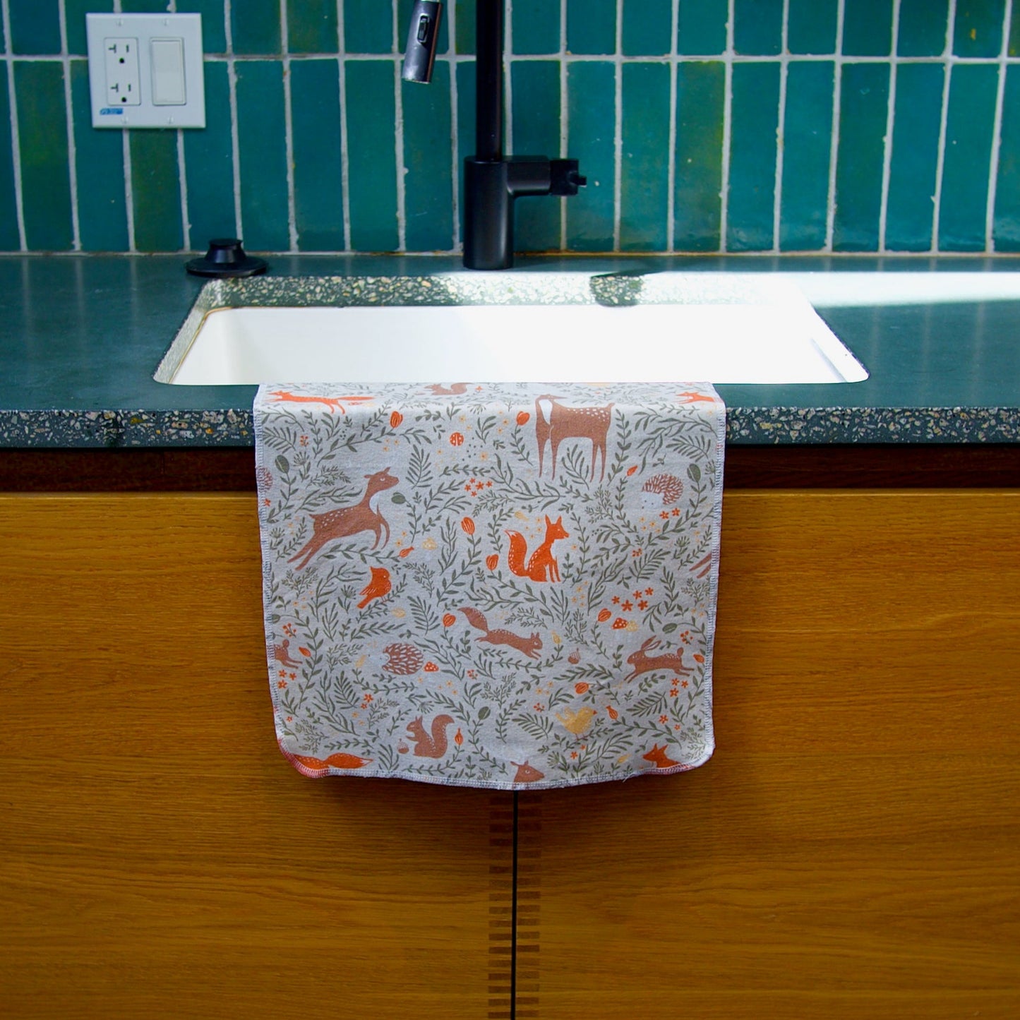Kitchen sink with an autumn woodland towel hanging over it, against a green tiled wall.
