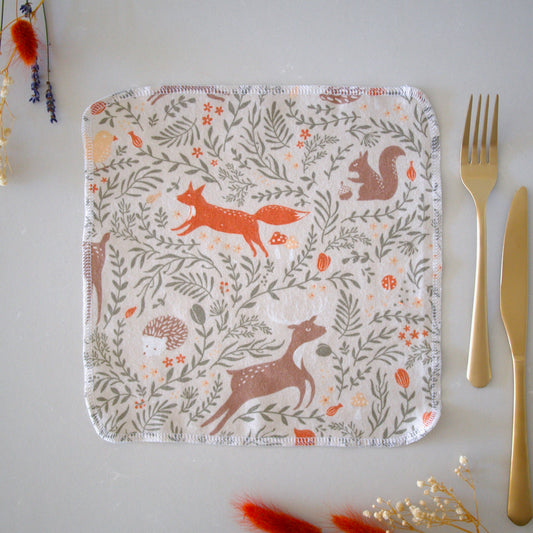 Eco-friendly napkin in an autumn woodland print featuring deer foxes squirrels and hedgehogs on a table with a gold fork and knife next to it