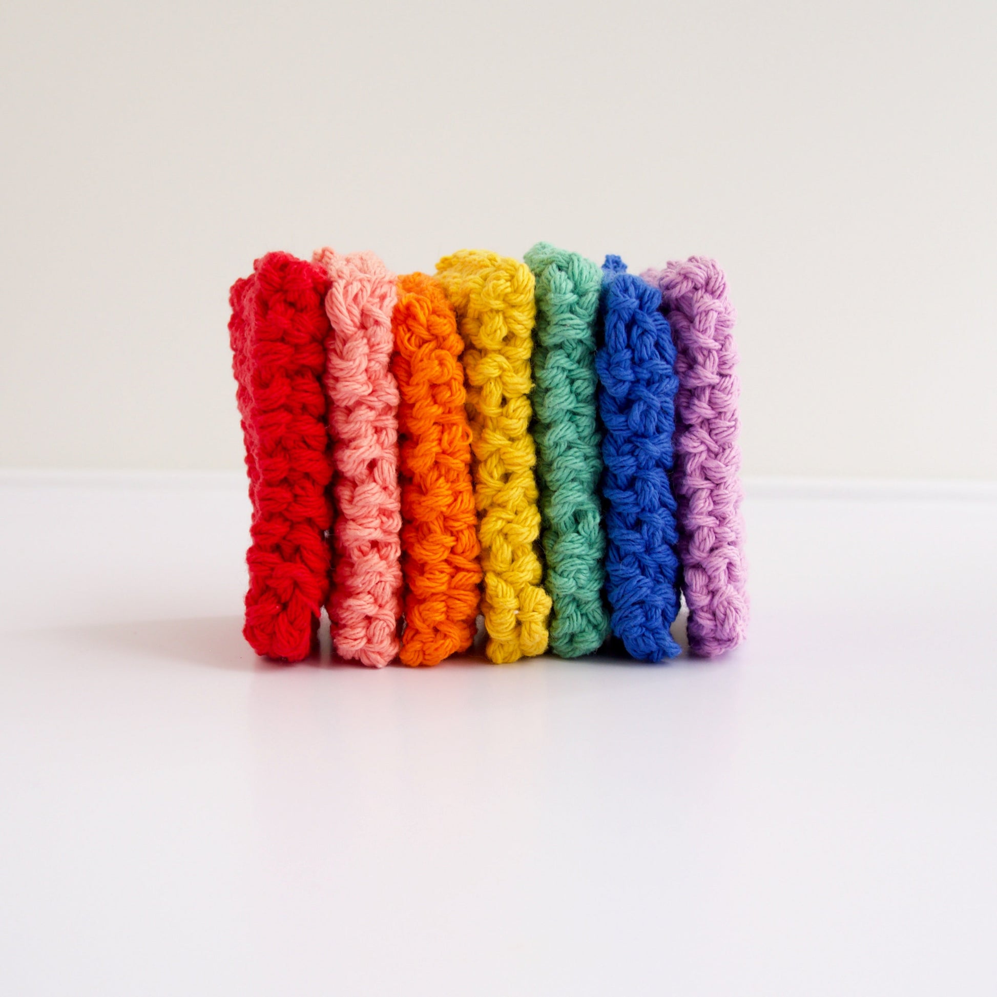 7 reusable cotton sponges lined up in a rainbow of colors from left to right, red, pink, orange, yellow, green, blue, light purple